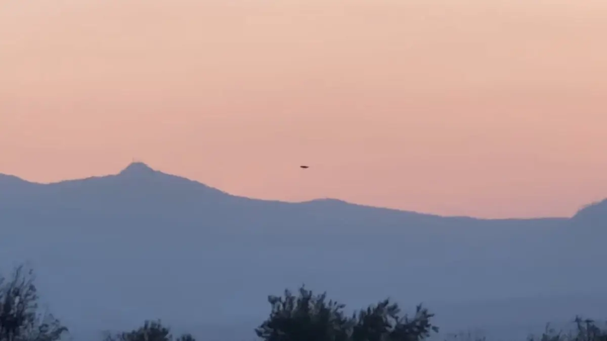 Misterioso ponto escuro pairando sobre as montanhas misteriosas Captura de tela granulada e de baixa resolução mostrando um suposto Fenômeno Aéreo Não Identificado (UAP) em forma de ponto escuro no céu de crepúsculo rosa pálido acima de silhuetas de montanhas borradas.